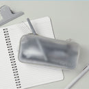 A translucent gray zippered pencil case with the brand name SKELT embossed on the front, styled on top of an open spiral notebook with a pen and clip.