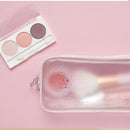 A translucent pink makeup pouch with condensation droplets, containing a pink makeup brush, shown next to a three-color eyeshadow palette on a solid pink background.