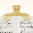 A translucent Sun-Star Ukanmuri Book Clip holding open a book with printed sheet music, photographed against a clean white background.