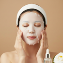 A woman with a white headband applies a translucent sheet mask to her face, with a skincare dropper bottle and a white flower in the foreground against a warm beige background.